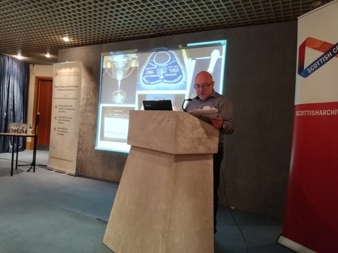 Harvey Kaplan at the CAHG 2019 Conference.. Harvey Kaplan at the CAHG 2019 Conference.. A person stands behind a lectern gesticulating, a powerpoint is behind them.