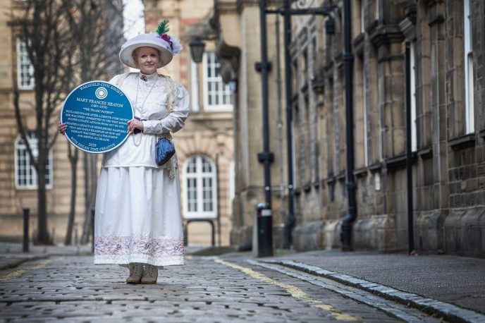 Sarah Cobham, in period costume, holding a blue plaque, near Wakefield town hall, with the blue plaque dedicated to Mary Francis Heaton; West Riding Pauper Lunatic Asylum, in 1818 | Gary Carlton of the Observer