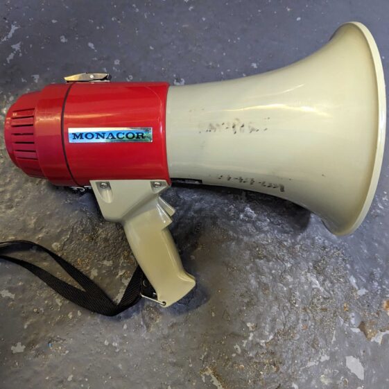 A red and white megaphone with faded pen writing on the white portion, resting on a grey background. | Image courtesy of Transport For All
