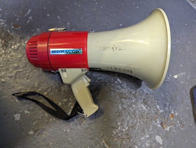 A red and white megaphone with faded pen writing on the white portion, resting on a grey background. | Image courtesy of Transport For All