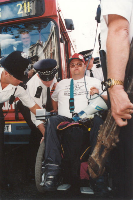 A man in a white t-shirt is lifted by the wheelchair by police. They are in front of a red London bus and the man has bungee cables and other things used to tie him to the bus on his lap | Image courtesy of Transport For All