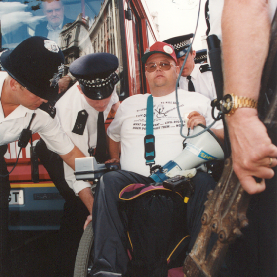 A man in a white t-shirt is lifted by the wheelchair by police. They are in front of a red London bus and the man has bungee cables and other things used to tie him to the bus on his lap | Image courtesy of Transport For All