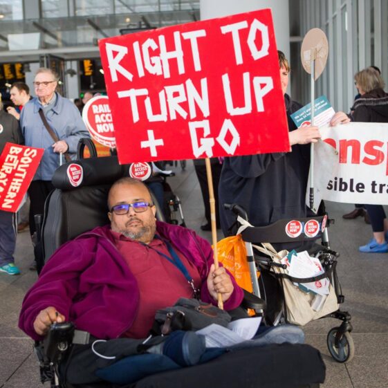 A man using a powerchair holds up a red protest sign that reads 